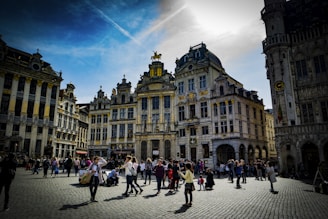 A bustling town square surrounded by ornate, historic buildings with intricate architectural details. People are casually walking, taking photos, and socializing under a partly cloudy sky. The sunlight highlights the golden accents on the structures, creating a lively and inviting atmosphere.