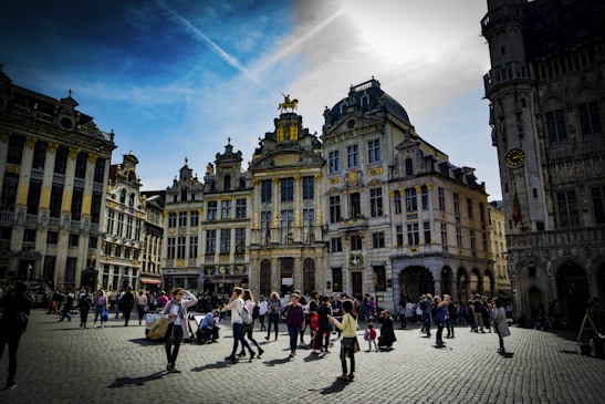 A bustling town square surrounded by ornate, historic buildings with intricate architectural details. People are casually walking, taking photos, and socializing under a partly cloudy sky. The sunlight highlights the golden accents on the structures, creating a lively and inviting atmosphere.