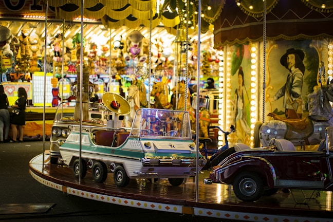 A vibrant amusement park scene featuring a carousel with vintage-style vehicles and horses. The background is filled with bright lights, stuffed animals, and colorful games, contributing to a lively carnival atmosphere.