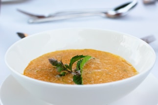 A bowl of warm soup with a smooth, orange-colored texture is garnished with fresh green herbs. The bowl is placed on a white saucer, set against a softly blurred background with silver cutlery visible, suggesting a dining setting.