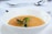 A bowl of warm soup with a smooth, orange-colored texture is garnished with fresh green herbs. The bowl is placed on a white saucer, set against a softly blurred background with silver cutlery visible, suggesting a dining setting.