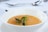 A bowl of warm soup with a smooth, orange-colored texture is garnished with fresh green herbs. The bowl is placed on a white saucer, set against a softly blurred background with silver cutlery visible, suggesting a dining setting.