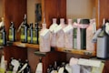 Several shelves display a variety of olive oil bottles. The bottles are in dark and light colors, with some wrapped in decorative packaging with tags. The shelves are wooden, and the setting appears to be a specialty or gourmet shop.
