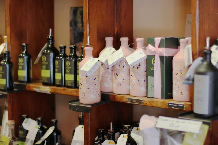 Several shelves display a variety of olive oil bottles. The bottles are in dark and light colors, with some wrapped in decorative packaging with tags. The shelves are wooden, and the setting appears to be a specialty or gourmet shop.