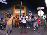 A street band is performing on a sidewalk, with several musicians playing brass instruments like trumpets and trombones. The group appears lively, with bystanders watching and enjoying the performance. The scene is colorful with lively signage in the background.