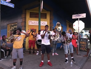 A street band is performing on a sidewalk, with several musicians playing brass instruments like trumpets and trombones. The group appears lively, with bystanders watching and enjoying the performance. The scene is colorful with lively signage in the background.