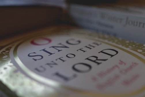 A close-up view of a book cover with the title 'Sing Unto the Lord'. The cover features elegant lettering surrounded by a decorative border with a muted color palette. The background includes blurred images of other books, suggesting a cozy, literary setting.