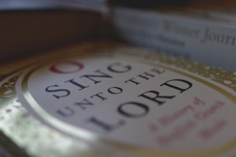 A close-up view of a book cover with the title 'Sing Unto the Lord'. The cover features elegant lettering surrounded by a decorative border with a muted color palette. The background includes blurred images of other books, suggesting a cozy, literary setting.