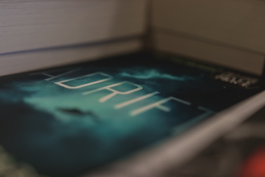 A close-up view of a book titled 'ADRIFT' with a mysterious, dark cover featuring a cloudy sky and a silhouette in the background. The book is placed between two other books, creating a sense of depth.