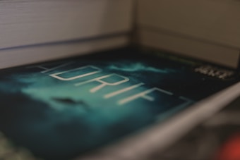 A close-up view of a book titled 'ADRIFT' with a mysterious, dark cover featuring a cloudy sky and a silhouette in the background. The book is placed between two other books, creating a sense of depth.