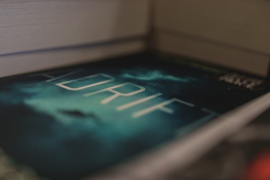 A close-up view of a book titled 'ADRIFT' with a mysterious, dark cover featuring a cloudy sky and a silhouette in the background. The book is placed between two other books, creating a sense of depth.