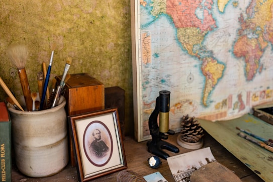 A wooden desk containing a variety of objects including art brushes in a ceramic holder, an old framed photograph, a vintage microscope, a pine cone, and a world map. The surface also has some old papers, a small rock, and a book titled 'FIELD CLUB FLORA'. The overall scene suggests an eclectic collection of items related to art, science, and travel.