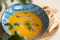 A vibrant orange soup garnished with seeds and herbs is presented in a patterned blue bowl. Next to the bowl, slices of brown bread are placed on a wooden board, adding a rustic touch. The setting includes a soft-focus view of green leaves in the foreground.