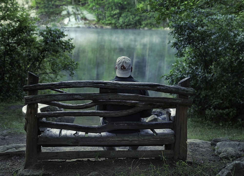 man sitting on brown wooden bench in front of body of water near trees at daytime