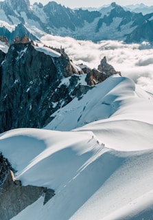 A group of mountaineers is trekking across a snowy ridge, surrounded by towering jagged peaks and fluffy white clouds. The light casts long shadows, adding depth to the pristine, untouched snow.