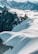 A group of mountaineers is trekking across a snowy ridge, surrounded by towering jagged peaks and fluffy white clouds. The light casts long shadows, adding depth to the pristine, untouched snow.