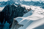 A breathtaking view of rugged mountain peaks with snow-covered slopes and a line of hikers trekking along a narrow ridge. The sky is partly cloudy, and the low-lying clouds create a dramatic contrast with the dark rock formations.