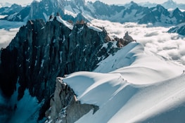 A breathtaking view of rugged mountain peaks with snow-covered slopes and a line of hikers trekking along a narrow ridge. The sky is partly cloudy, and the low-lying clouds create a dramatic contrast with the dark rock formations.