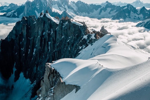 A breathtaking view of rugged mountain peaks with snow-covered slopes and a line of hikers trekking along a narrow ridge. The sky is partly cloudy, and the low-lying clouds create a dramatic contrast with the dark rock formations.