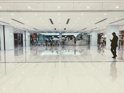 A spacious and modern indoor shopping area with bright lighting and shiny, reflective floors. People are casually walking and standing around, some interacting with their phones. There are stores and seating areas visible in the background.