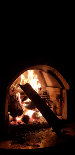A cozy stonehouse fireplace glowing warmly with stacked logs beside it.