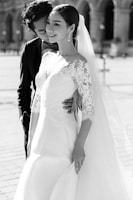 A couple embraces with the woman wearing an elegant lace wedding dress and veil. The man is dressed in a dark suit, gently embracing the woman from behind. Their expressions convey happiness and affection, set against an outdoor background with soft focus.