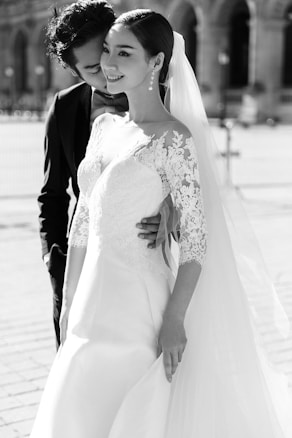 A couple embraces with the woman wearing an elegant lace wedding dress and veil. The man is dressed in a dark suit, gently embracing the woman from behind. Their expressions convey happiness and affection, set against an outdoor background with soft focus.