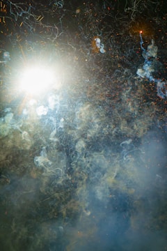A burst of bright light and swirling smoke fills the scene, with intricate patterns of light trails and sparks scattered throughout. The background is a dark, cloudy sky, creating a dramatic contrast with the luminous elements.