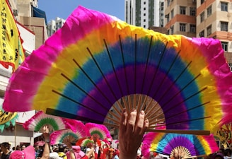 Colorful fans are held up by a crowd, creating a vibrant display against the backdrop of city buildings. Bright shades of pink, yellow, blue, and green dominate the fan designs. In the background, banners with Chinese characters and participants in pink hats suggest a festive or cultural event.