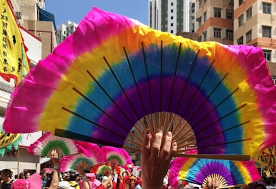 Colorful fans are held up by a crowd, creating a vibrant display against the backdrop of city buildings. Bright shades of pink, yellow, blue, and green dominate the fan designs. In the background, banners with Chinese characters and participants in pink hats suggest a festive or cultural event.