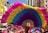 Colorful fans are held up by a crowd, creating a vibrant display against the backdrop of city buildings. Bright shades of pink, yellow, blue, and green dominate the fan designs. In the background, banners with Chinese characters and participants in pink hats suggest a festive or cultural event.