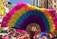 Colorful fans are held up by a crowd, creating a vibrant display against the backdrop of city buildings. Bright shades of pink, yellow, blue, and green dominate the fan designs. In the background, banners with Chinese characters and participants in pink hats suggest a festive or cultural event.