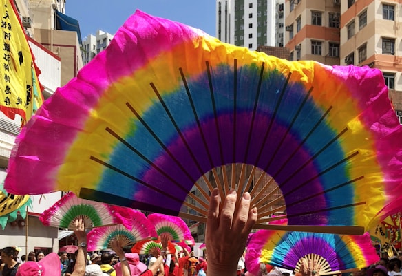Colorful fans are held up by a crowd, creating a vibrant display against the backdrop of city buildings. Bright shades of pink, yellow, blue, and green dominate the fan designs. In the background, banners with Chinese characters and participants in pink hats suggest a festive or cultural event.
