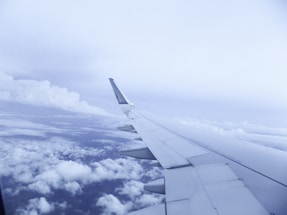 A view of an airplane wing in flight depicted against a backdrop of clouds and a soft blue sky. The wing occupies a significant portion of the frame, emphasizing the experience of air travel.