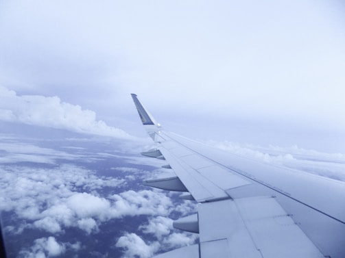 A view of an airplane wing in flight depicted against a backdrop of clouds and a soft blue sky. The wing occupies a significant portion of the frame, emphasizing the experience of air travel.