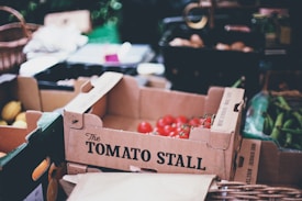 A cardboard box labeled 'The Tomato Stall' contains several ripe red tomatoes. Surrounding the box are other types of vegetables and more cardboard boxes. There are also wicker baskets and some blurred market produce in the background.
