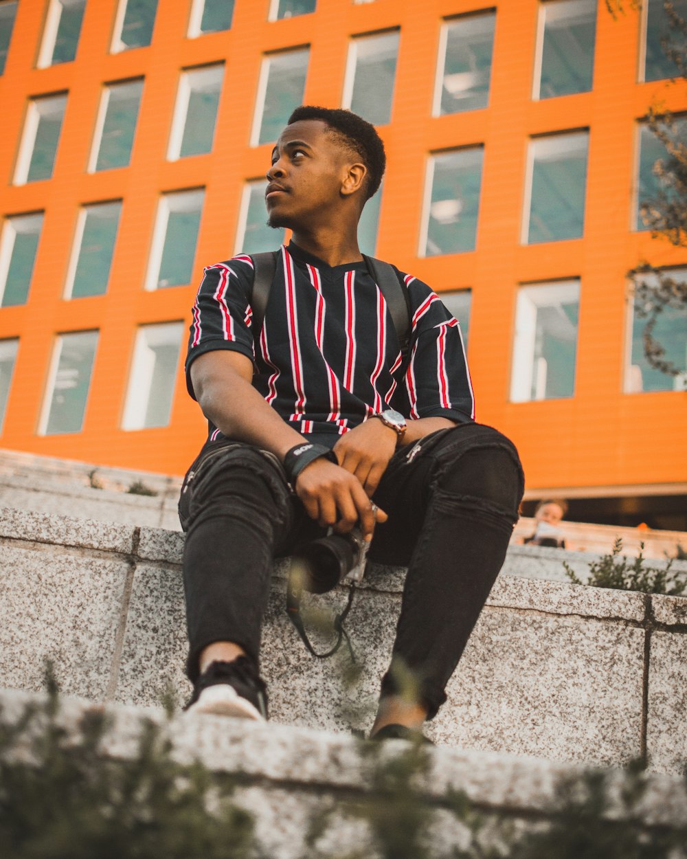 man wearing black and red striped V-neck shirt sitting on concrete stairs looking sideways during daytime