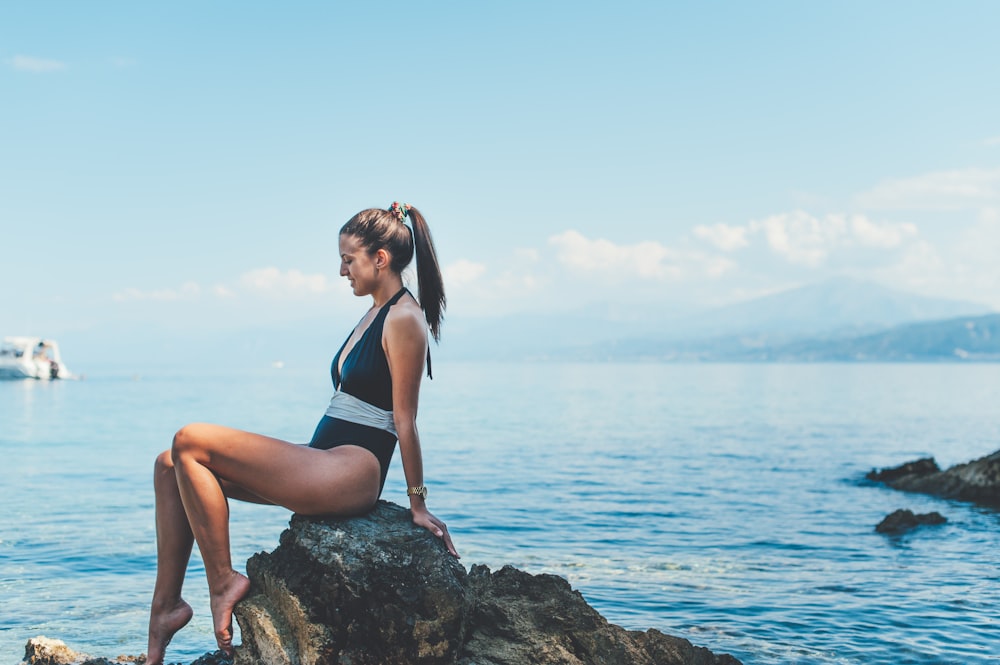Donna che indossa monokini seduta su roccia marrone vicino alla riva