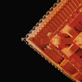 A close-up view of a traditional architectural structure, featuring intricate wooden beams and details painted in a vibrant orange, set against a stark black background. The image captures the geometric precision and craftsmanship of the design.