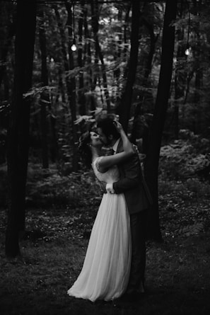 A couple embraces and kisses in a forest setting, surrounded by tall trees with sparse leaves. The scene appears romantic and intimate, accentuated by the black and white color palette, creating a timeless and classic atmosphere.