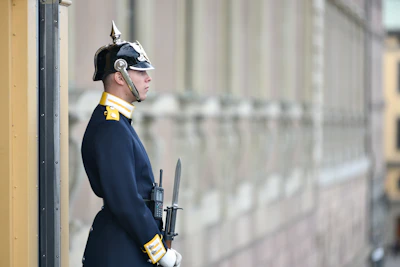 A uniformed guard stands at attention beside a building. The guard wears a dark blue uniform with gold accents and a shiny helmet with a spike on top. The guard holds a rifle with a bayonet attached, and appears to be standing in a formal posture.