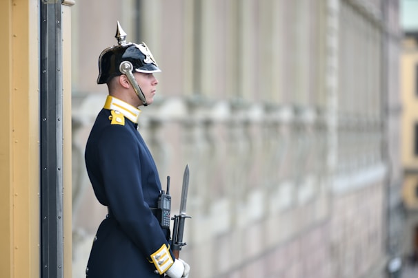 A uniformed guard stands at attention beside a building. The guard wears a dark blue uniform with gold accents and a shiny helmet with a spike on top. The guard holds a rifle with a bayonet attached, and appears to be standing in a formal posture.