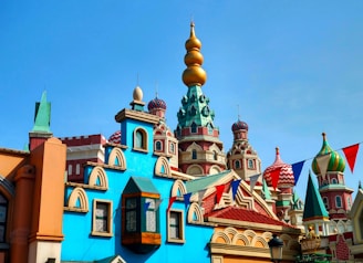 A vibrant architectural scene featuring colorful onion domes and intricate building designs reminiscent of Russian architecture. The sky is clear, highlighting the vivid colors and patterns on the structures. Strings of flags add a festive atmosphere.