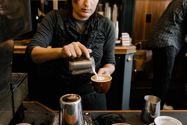 A person is pouring steamed milk into a coffee cup to create latte art in a cozy coffee shop environment. The individual is focused on the task, and the setting includes various coffee-making equipment and a warm, wooden interior.