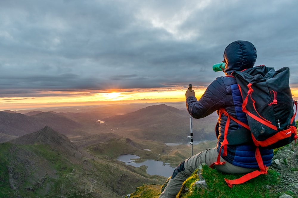 man sitting on cliffhanger looking at mountain under gloomy clouds