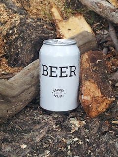 A white can labeled 'BEER' with 'Garage Project' is set on a rustic, outdoor background. Surrounding the can are pieces of bark, wood, and soil, giving a natural and earthy vibe.