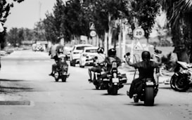 Several motorcyclists ride down a tree-lined street, with cars and traffic signs in the background. The image is black and white, highlighting the contrast of the bikers against the road.