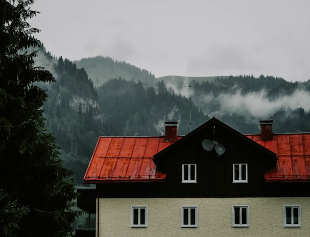 A house with a striking red roof is positioned against a backdrop of misty, forested mountains. The mist partially shrouds the evergreen trees, creating a sense of depth and mystery. Two satellite dishes are visible on the side of the house, suggesting modern connectivity amidst a rural setting.
