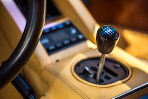 A close-up view of a manual gear shift knob inside a car. The gear knob has a black finish with white numbers and a diagram representing the gear positions. The surrounding interior is upholstered in tan leather with visible stitching.
