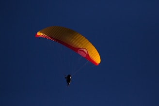 A lone paraglider with a vibrant orange and red canopy is soaring against a deep blue sky. The silhouette of the pilot is visible beneath the parachute, creating a sense of freedom and adventure.
